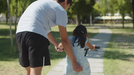 rear view of father teaching his daughter to ride bike in park