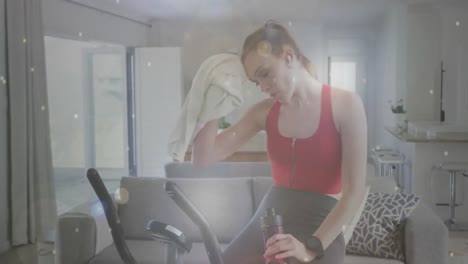 Animation-of-white-spots-over-caucasian-woman-exercising,-using-stationary-bike-at-home