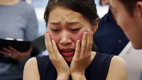 a woman appears overwhelmed in the workplace, covering her face with her hands as emotions take over. colleagues can be seen in the background, engaged in their tasks.