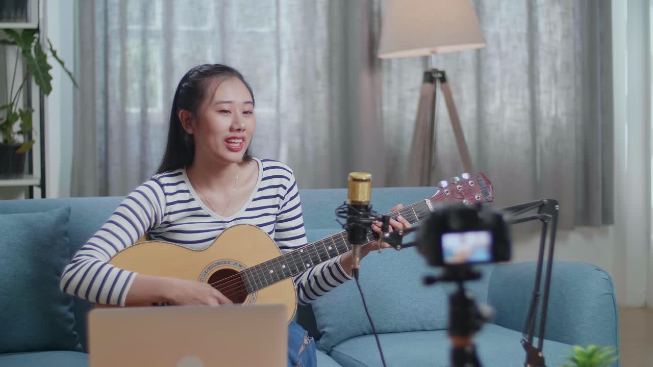 mujer asiática con una computadora portátil cantando y tocando una guitarra mientras transmite en vivo en casa
