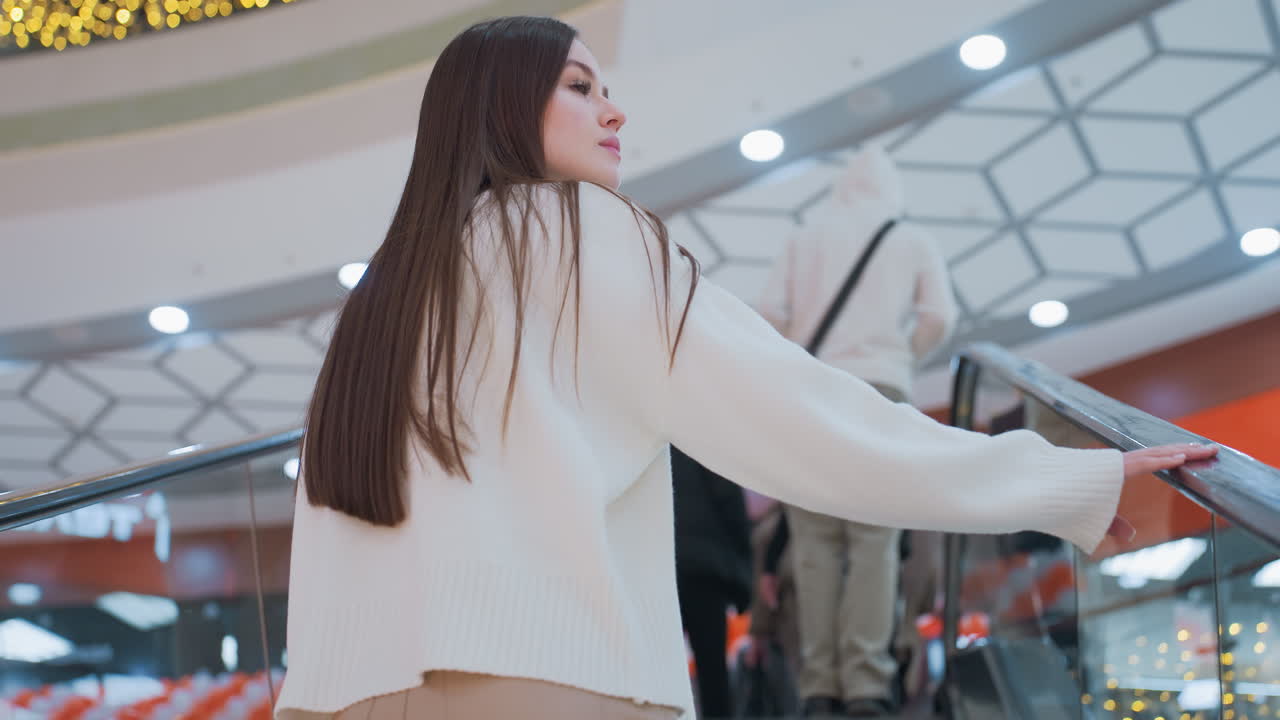 Rear view of woman in white sweater with long brown hair ascending escalator in shopping mall, she gently places hand on rail while moving up, with other people ahead
