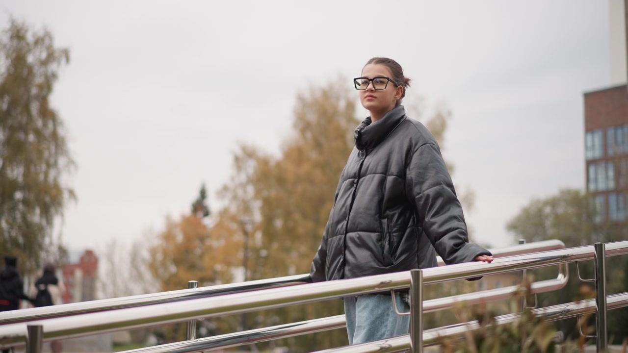 Close up of young woman wearing glasses and black puffer jacket standing on outdoor ramp with metal railings. Woman maintains same pose throughout looking around in different directions