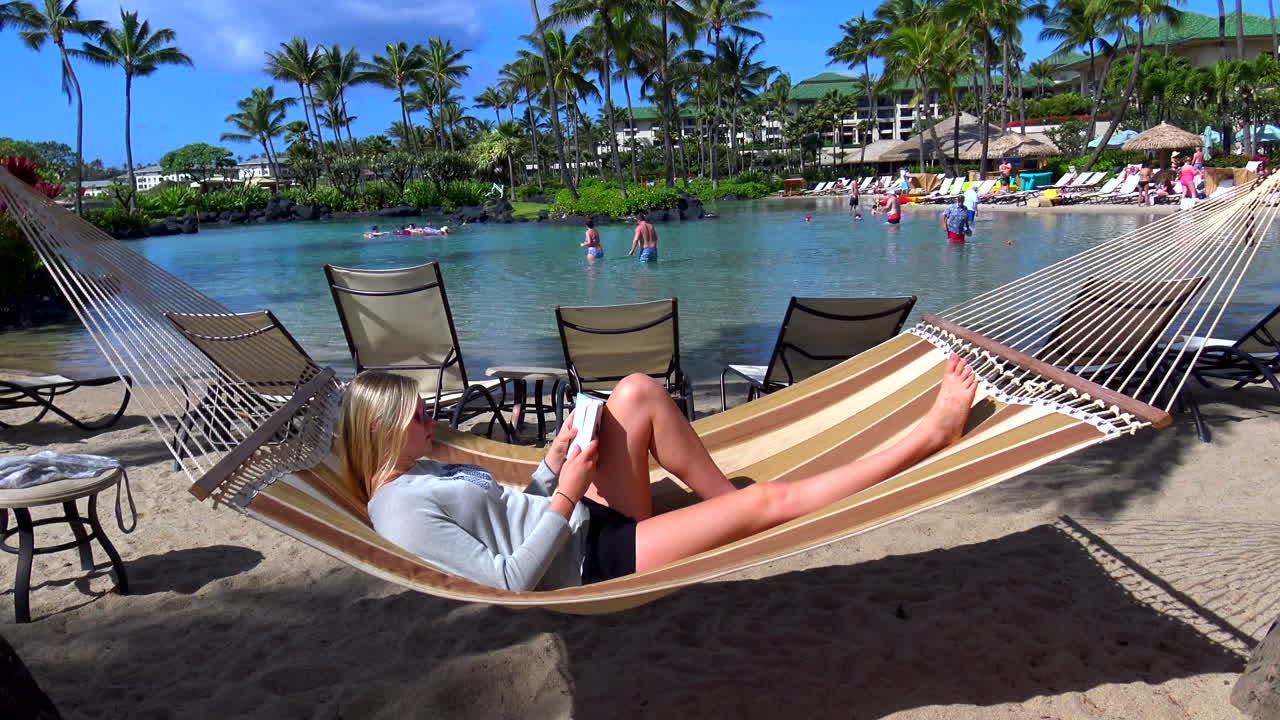 joven mujer rubia relajándose en una hamaca en la piscina del hotel resort y laguna de natación en la playa de poipu, kauai, hawaii, 4k, estática, leyendo un libro