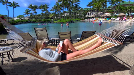 Joven-Mujer-Rubia-Relajándose-En-Una-Hamaca-En-La-Piscina-Del-Hotel-Resort-Y-Laguna-De-Natación-En-La-Playa-De-Poipu,-Kauai,-Hawaii,-4k,-Estática,-Leyendo-Un-Libro