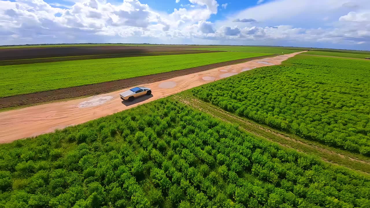 Following Tesla Cybertruck moving by the road near the green farmland. Futuristic automobile in the beautiful nature landscape. FPV drone footage.