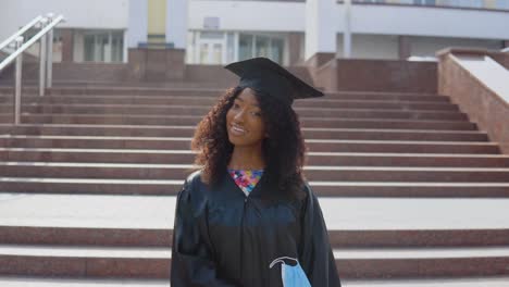 a young african american female graduate stands in front of the camera and removes a protective medical mask from her face. the student stands on the stairs outside.