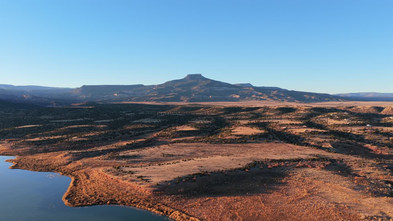 Morning sunlight hits the volcanic basalt peak of Cerro Pedernal above Abiquiu Lake. The high desert plateau landscape features arid soil and a rippled blue reservoir in northern New Mexico