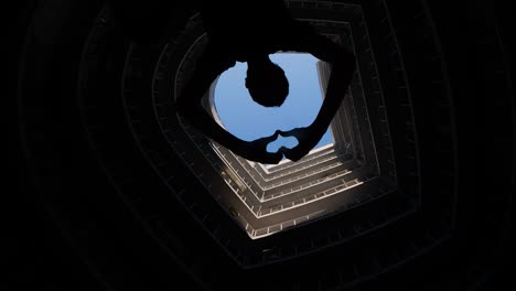 modern round building with blue sky on top. silhouette of a man who shoots video on a phone or smartphone. young man making a heart shape with his hands. male showing a heart shaped. feelings emotions