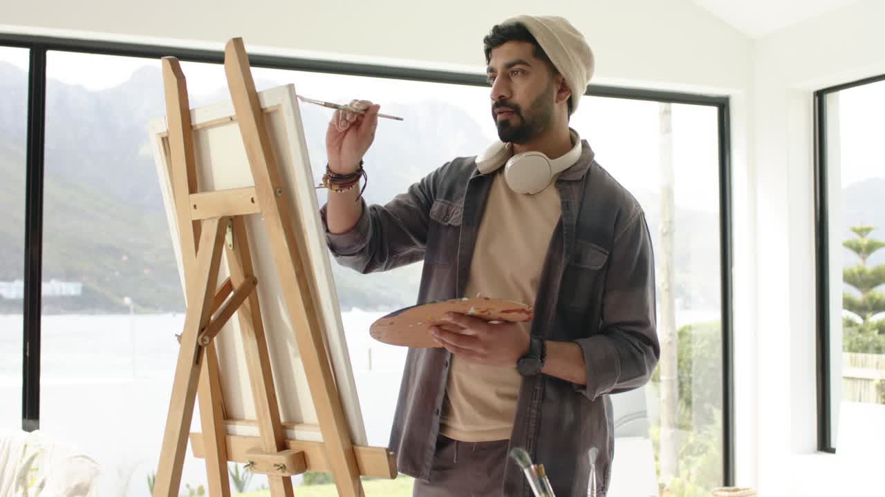 Dopo aver esaminato le aree di lavoro, uomo indiano che intinge il pennello e dipinge alla finestra di uno studio, ispirato dal lago