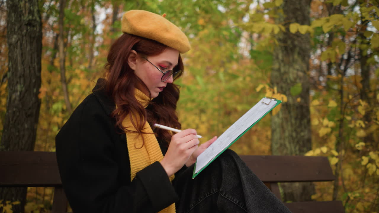 Artist in yellow beret and muffler deeply focused on her drawing seated on a bench surrounded by autumn leaves, embodying artistic concentration in a natural setting