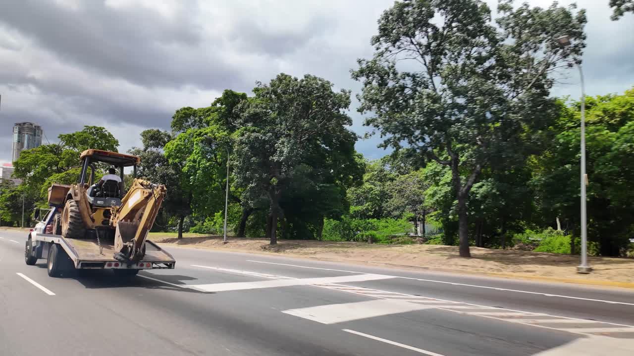 초록색 나무와 구름이 낀 하늘로 둘러싸인 고속도로를 달리는 평판 트럭에 실려 운반되는 노란색 백호 로더