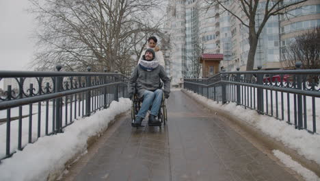 Vorderansicht-Einer-Muslimischen-Frau,-Die-Ihren-Behinderten-Freund-Im-Rollstuhl-Im-Winter-1-Auf-Einem-Spaziergang-In-Der-Stadt-Mitnimmt
