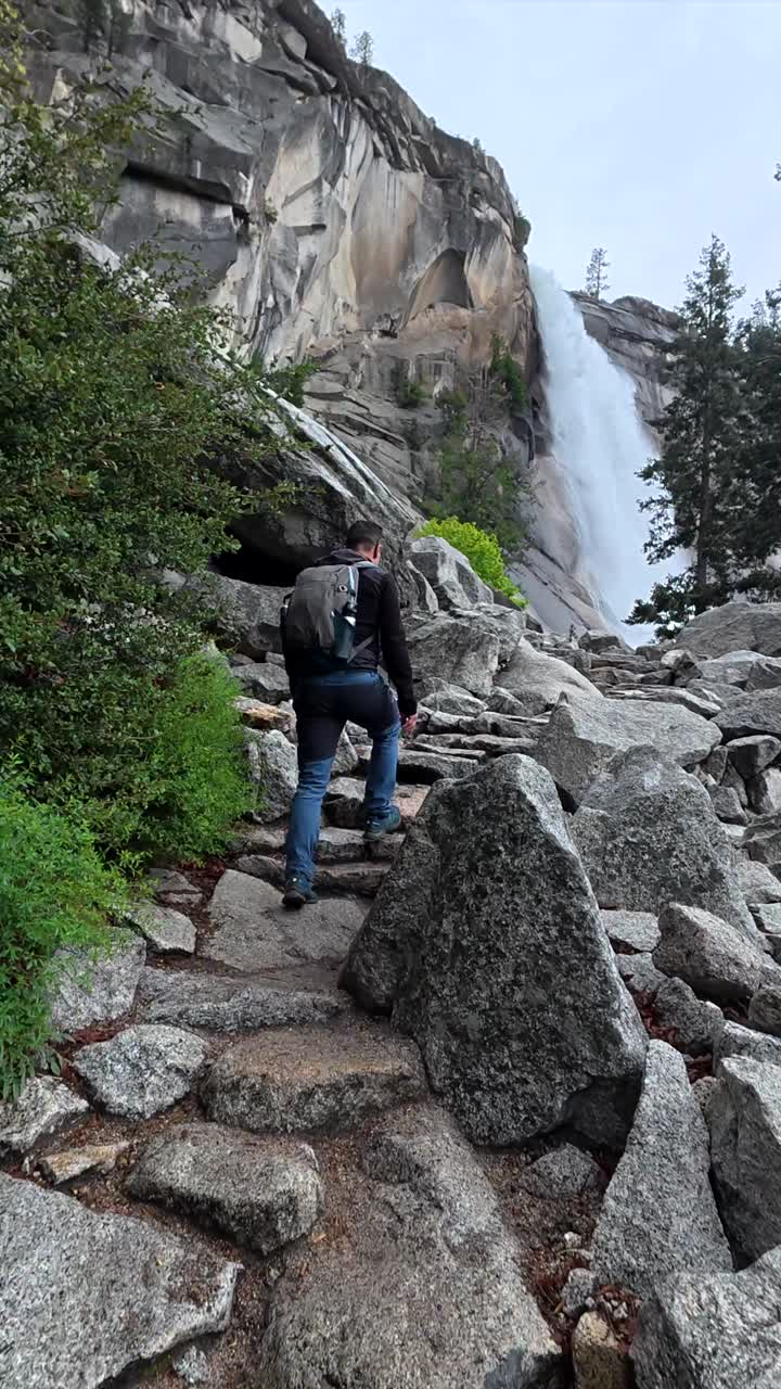 Uomo in escursione su sentiero roccioso verso una cascata maestosa