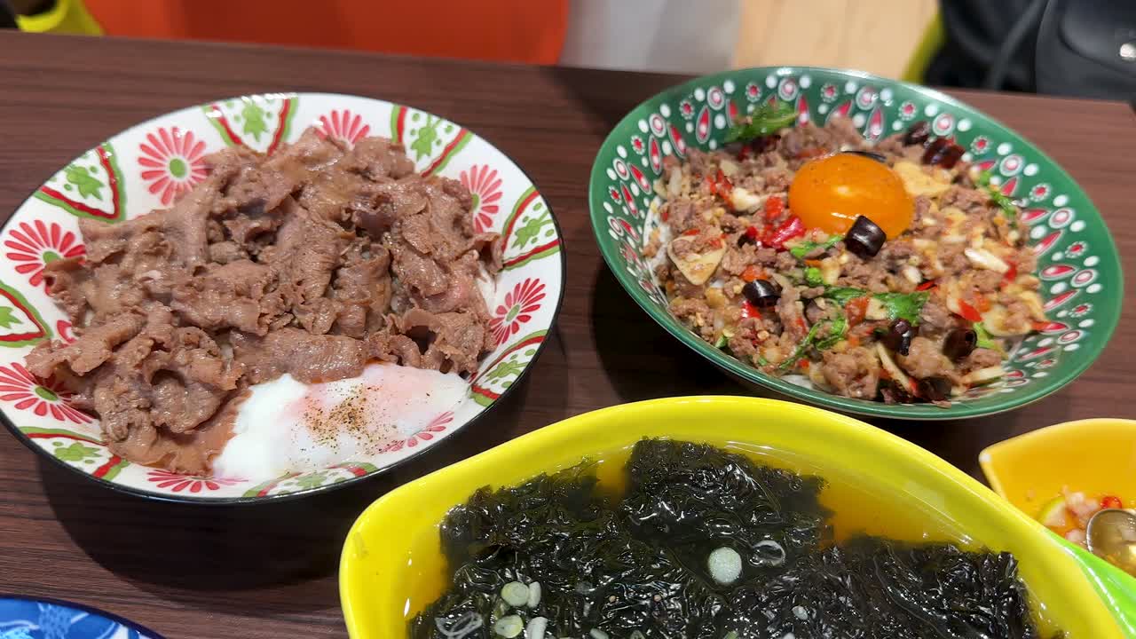 A vibrant overhead sequence of assorted Asian rice bowls with beef, shrimp, egg, vegetables, and seaweed soup, under bright indoor lighting with smooth camera panning