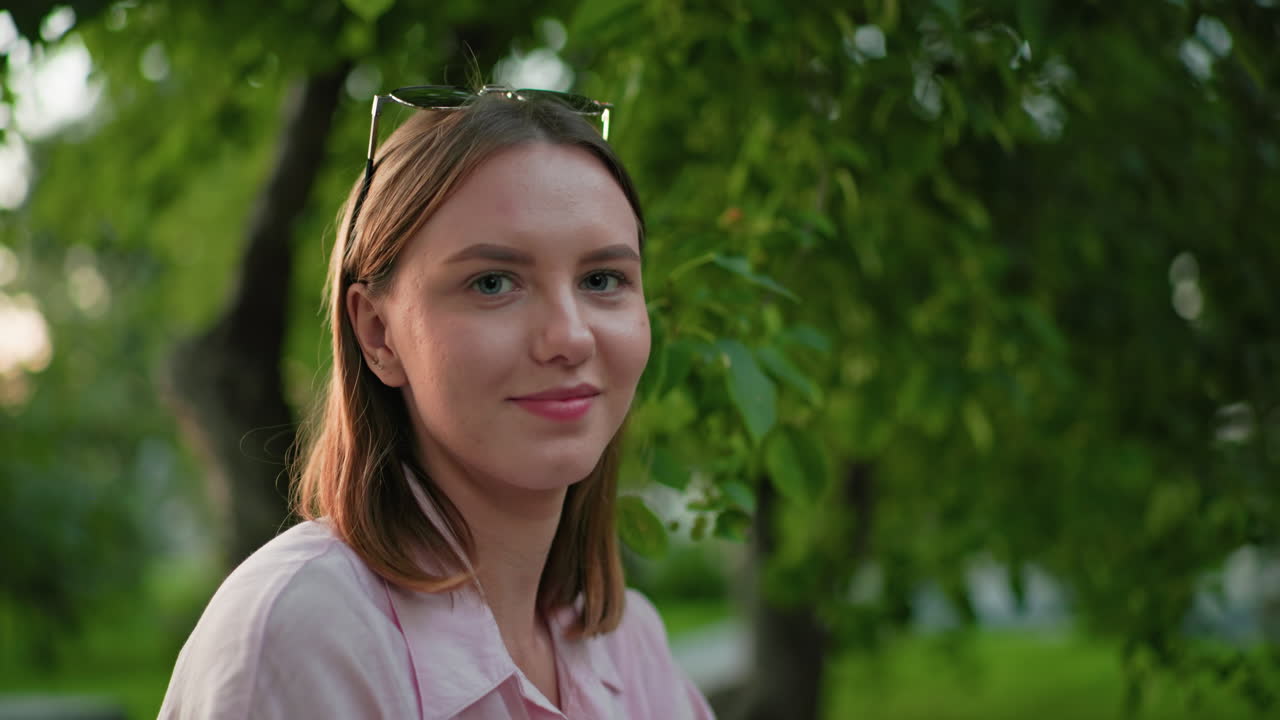 따뜻한 미소를 짓는 여성의 클로즈업, 울창한 녹색 나무와 부드러운 야외 조명으로 둘러싸여 평온하고 긍정적인 분위기를 만니다.