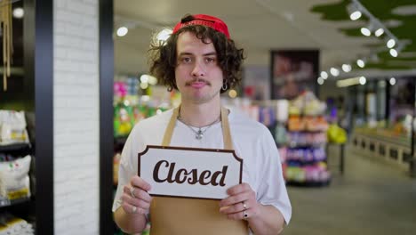 Sad-guy-supermarket-worker-with-curly-hair-holds-in-his-hands-a-sign-with-the-inscription-Closed-while-in-a-closed-supermarket