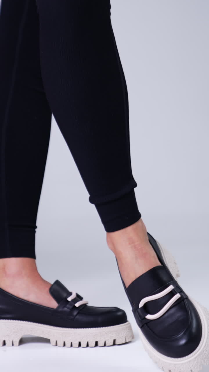 Unrecognized female in black pants wears modern shoes on white soles. Woman standing beside red chair demonstrating stylish footwear. White backdrop. Vertical video