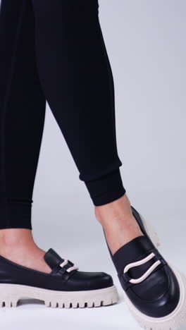 unrecognized female in black pants wears modern shoes on white soles. woman standing beside red chair demonstrating stylish footwear. white backdrop. vertical video