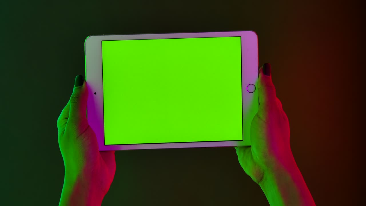 African American woman holds in her hand a white tablet with a green screen chroma key. Close up gadget in a horizontal position against a background of bright multicolored neon lights. Slow motion