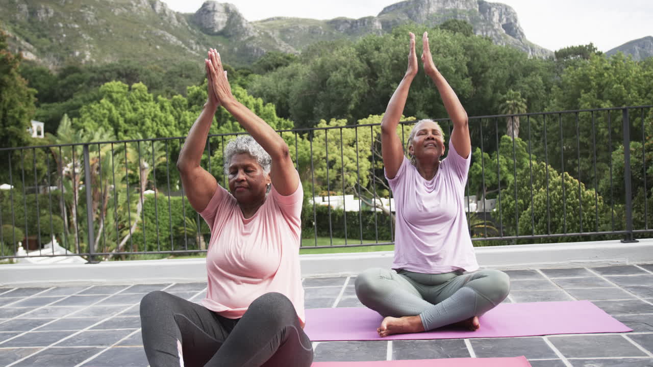 Senior african american female friends practicing yoga meditation sitting on balcony, slow motion