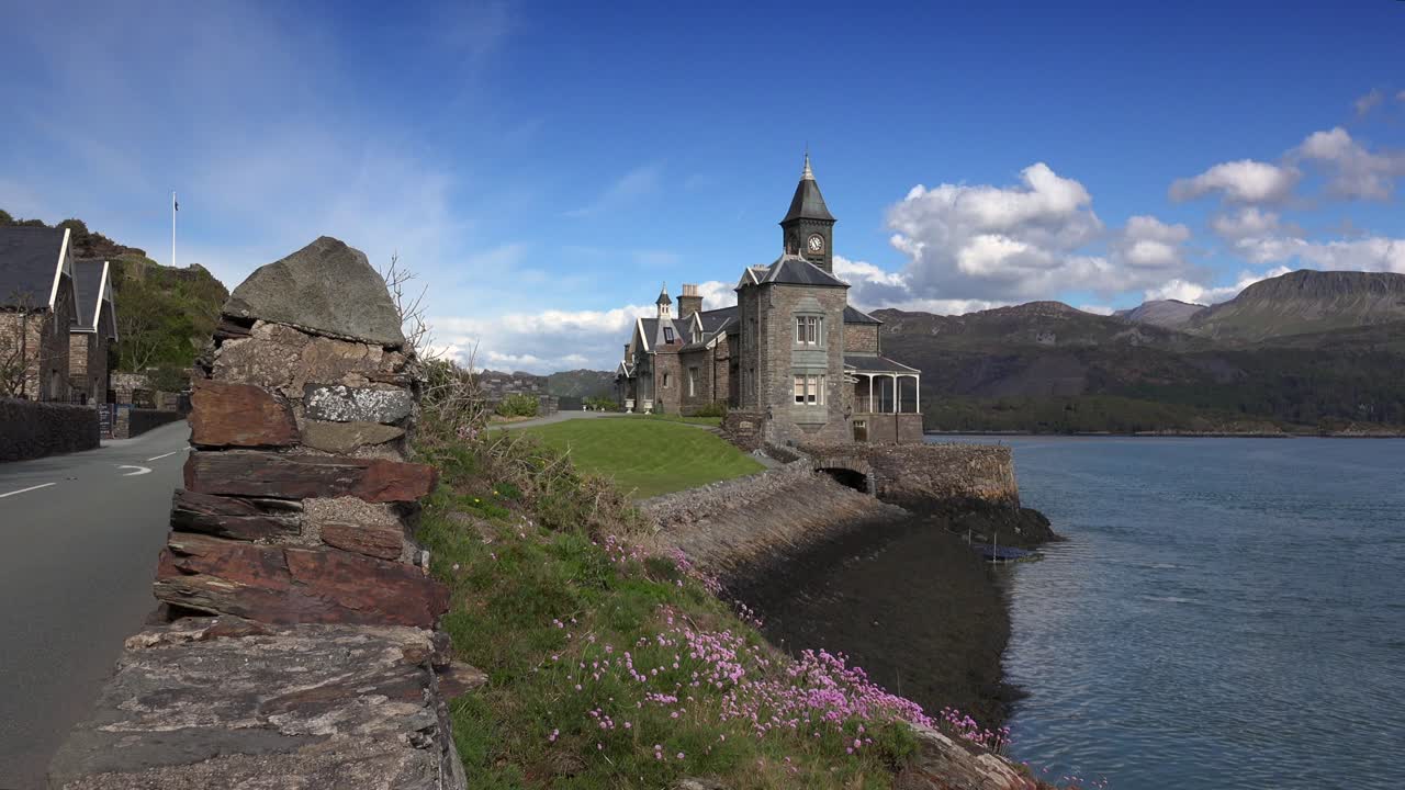 Scenic coastal view with a historic stone building by the water, a winding road, and mountains in the background under a blue sky with scattered clouds