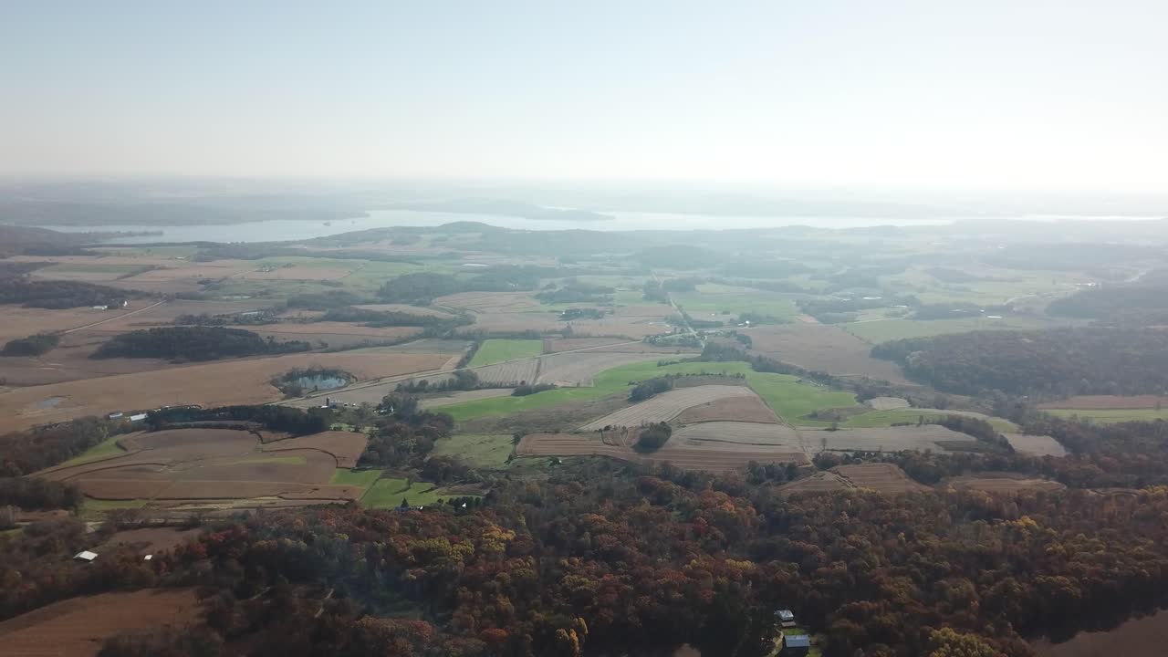 Aerial view over rolling farmland and patchy fields with Lake Wisconsin shimmering in the distance. Captures rural Midwest landscape under clear morning light.