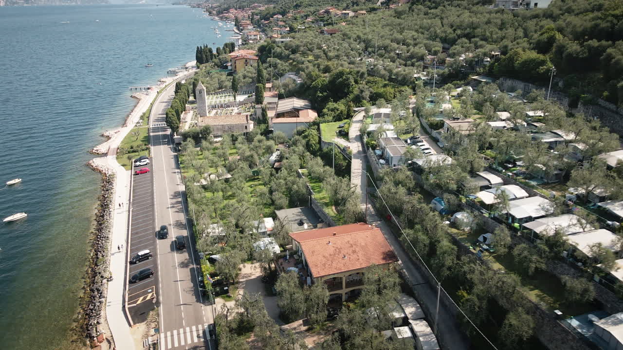 Göl kenarındaki bir şapel ve zeytinliklerden geri çekilerek kıyı yolunu, plajı ve masmavi suyu ortaya çıkaran, Garda Gölü kıyısı boyunca akıcı bir 4K drone geri çekimi.