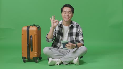 full body of asian male traveler with luggage looking at the passport, smiling, and showing okay gesture while sitting in the green screen background studio