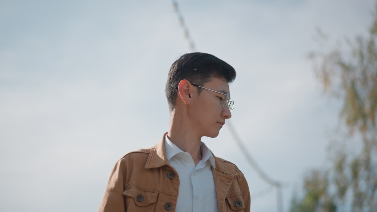 空を見上げる若いアジア人男性。柔らかな映画のような光が、ストリングライトと高い白樺の木々がある屋外の街並みを照らし出します。茶色のジャケットに透明な眼鏡、かすかなフィルムグレイン、ポートレートフレーミングの変化。