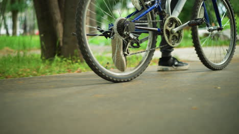 primer plano de alguien caminando al lado de una bicicleta a lo largo de una pasarela del parque, con fondo de hierba, los cordones de los zapatos flotan mientras se mueve, el enfoque está en las piernas y la bicicleta, con árboles en el fondo