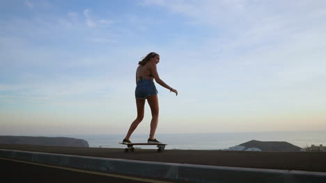 en medio de las montañas, una elegante joven patinadora monta su tabla en pantalones cortos en un camino de montaña al atardecer, toda la escena capturada en hipnótica cámara lenta