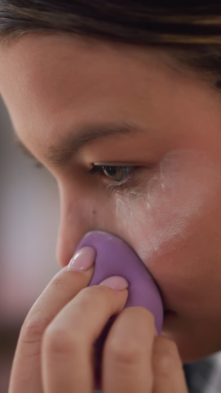 Primer plano de un adolescente aplicando polvos translúcidos con una esponja morada, movimiento decidido, ambiente de ensayo entre bastidores, preparándose para la actuación, expresión de seguridad, brillo de luces de estudio, práctica de creador de contenido