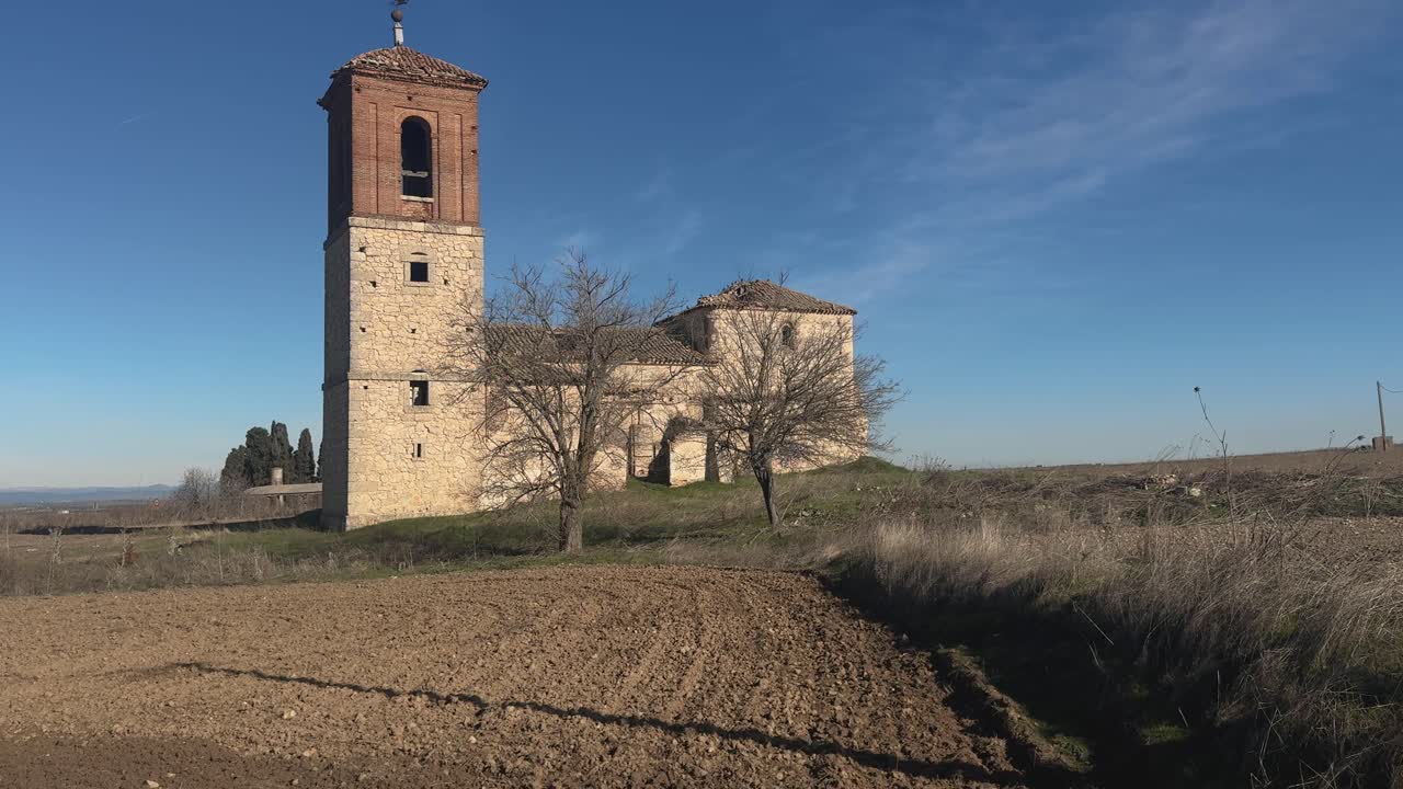 Inclinazione lenta verso l'alto a 24fps che mostra una chiesa in rovina con alberi spogli in primo piano e terreno arato, sotto un cielo azzurro e limpido