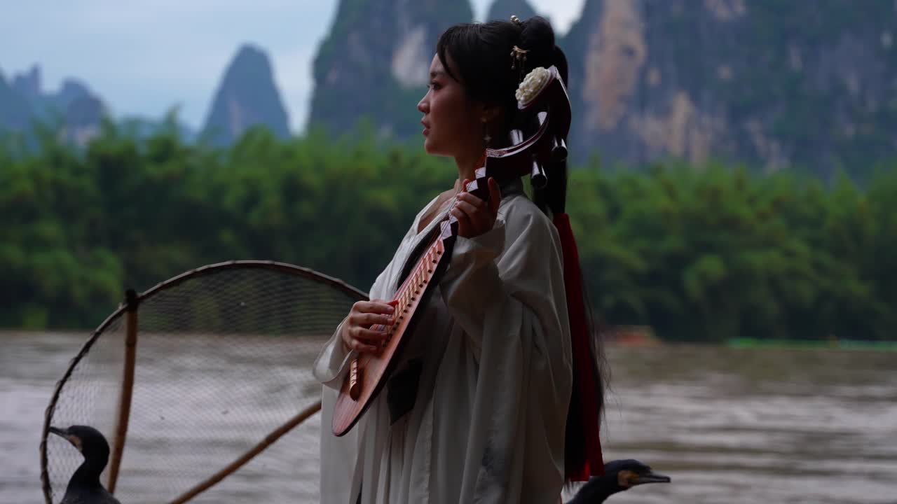 Hanfu Dynasty girl plays a pipe on a bamboo raft by Li River with scenic mountains in background