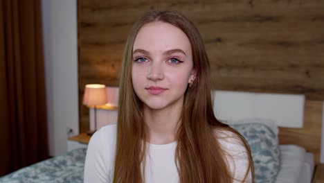 Smiling-pretty-young-woman-in-good-mood-looking-at-camera,-smiles-gently-on-bedroom-background