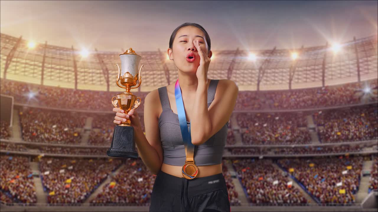 mujer asiática con una medalla de oro sosteniendo un trofeo de oro y gritando positivamente con la mano sobre la boca en el estadio