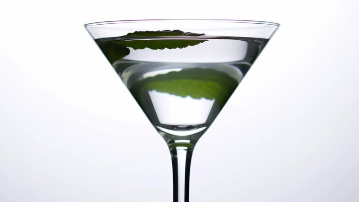 Movimento ravvicinato di un bicchiere da martini riempito con liquido trasparente mentre una singola foglia di menta galleggia, poi si posa vicino al bordo sotto una forte retroilluminazione ad alta chiave con riflessi morbidi, uno stile pulito e minimale, e