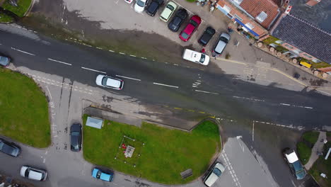 Top-Down-Luftdrohnenaufnahme-Von-Silbernen-Und-Roten-Autos-Und-Weißen-Lieferwagen,-Die-Auf-Einer-überfluteten-Straße-In-Einem-Kleinen-Britischen-Dorf-Durch-Wasser-Fahren