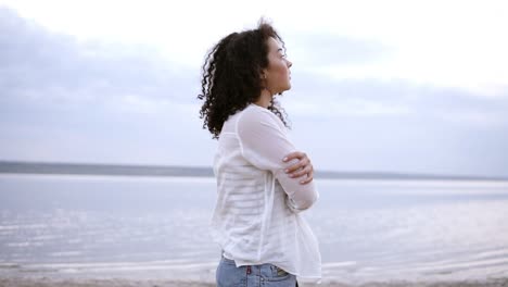 Close-up-side-footage-of-an-attractive-young-woman-walking-by-seaside-in-water,-wearing-a-white-shirt.-Curly-brunette-girl-in-cloudy-morning-walking-outdoors