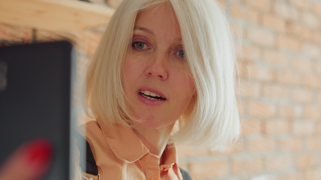 Closeup portrait of female florist in black apron with short blonde hair and orange shirt, focused expression while working in rustic workshop with brick wall background, natural light highlighting face