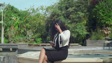a businesswoman typing on her phone outdoors with a pool in the background.
