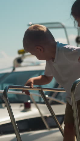 toddler boy looks down from yacht drawbridge landing with mother and sister in city port. junior brother scared to go down vessel ladder with family