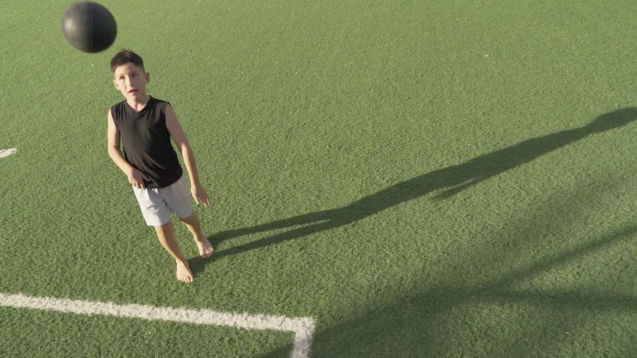 Talented barefoot boy playing with soccer ball on synthetic green field, enjoying an athletic activity under bright sunlight