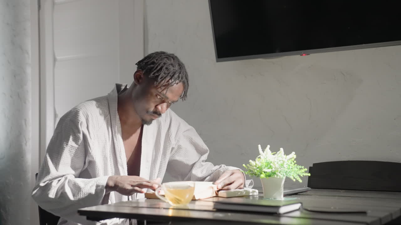 Man in white robe sitting at table, reading book and sipping tea. Soft light from window illuminates peaceful, modern living room, creating a calm atmosphere for relaxation and personal time