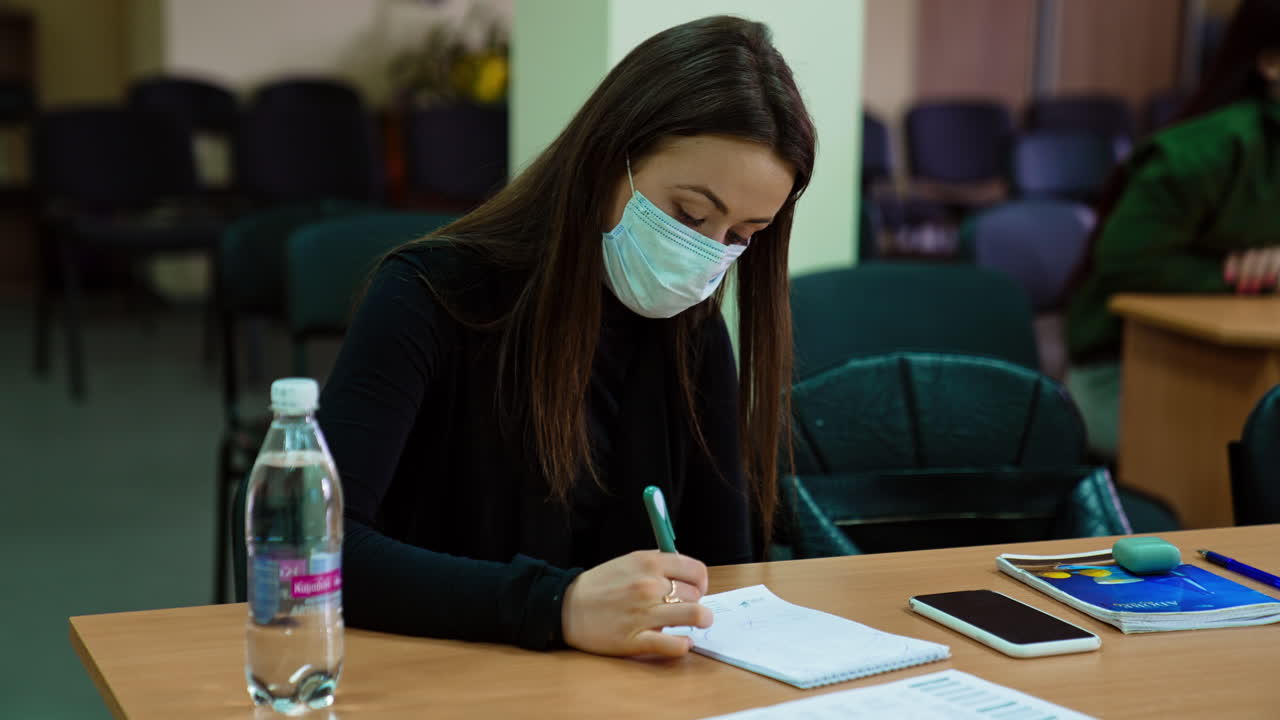 Long-haired brunette woman wearing mask sitting at the desk. Woman taking notes listening to the lecturer. Lots of students at backdrop.