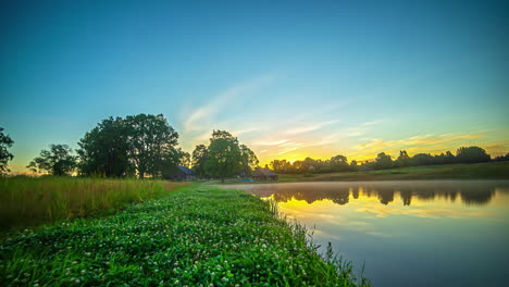 Verdant-riverbank-at-dawn-with-colorful-clouds-reflecting-on-river-water-surface
