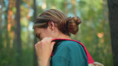 side view of woman adjusting strap of red backpack against background of lush forest with tall trees, with a calm expression and serene natural surroundings