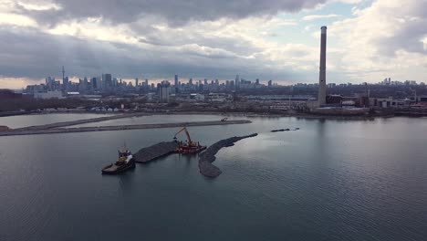 breakwater construction by excavator moving rocks from barge on water with toronto skyline