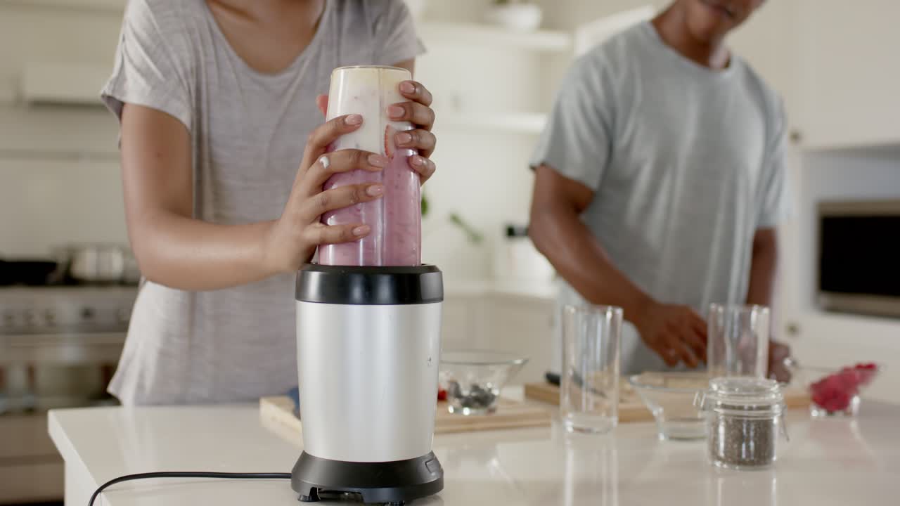Pareja afroamericana colocando un vaso en la base, encendiendo el motor, licuando una bebida rosa y sirviéndola en la isla de la cocina.