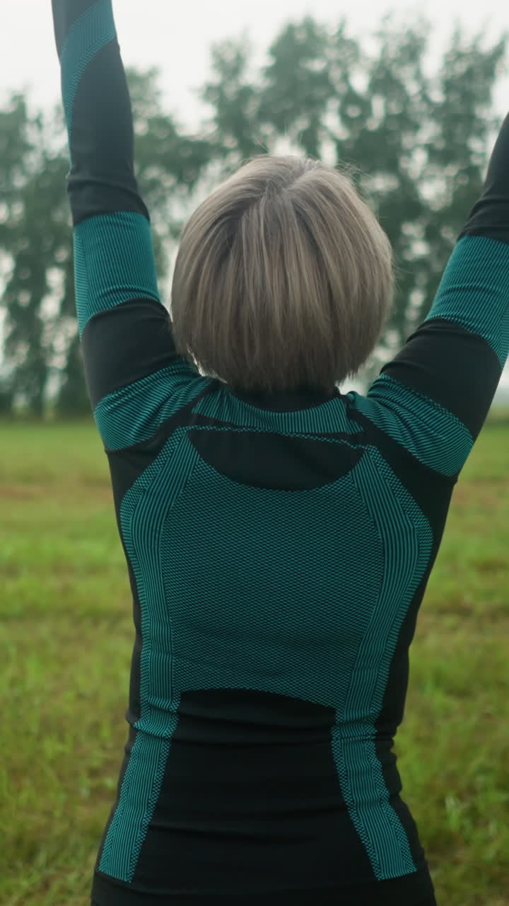 vista trasera de una mujer de cabello gris de pie con la cabeza ligeramente levantada, levantando los brazos en un movimiento sereno, practicando yoga en un vasto campo abierto bajo un cielo nublado con árboles borrosos en la distancia